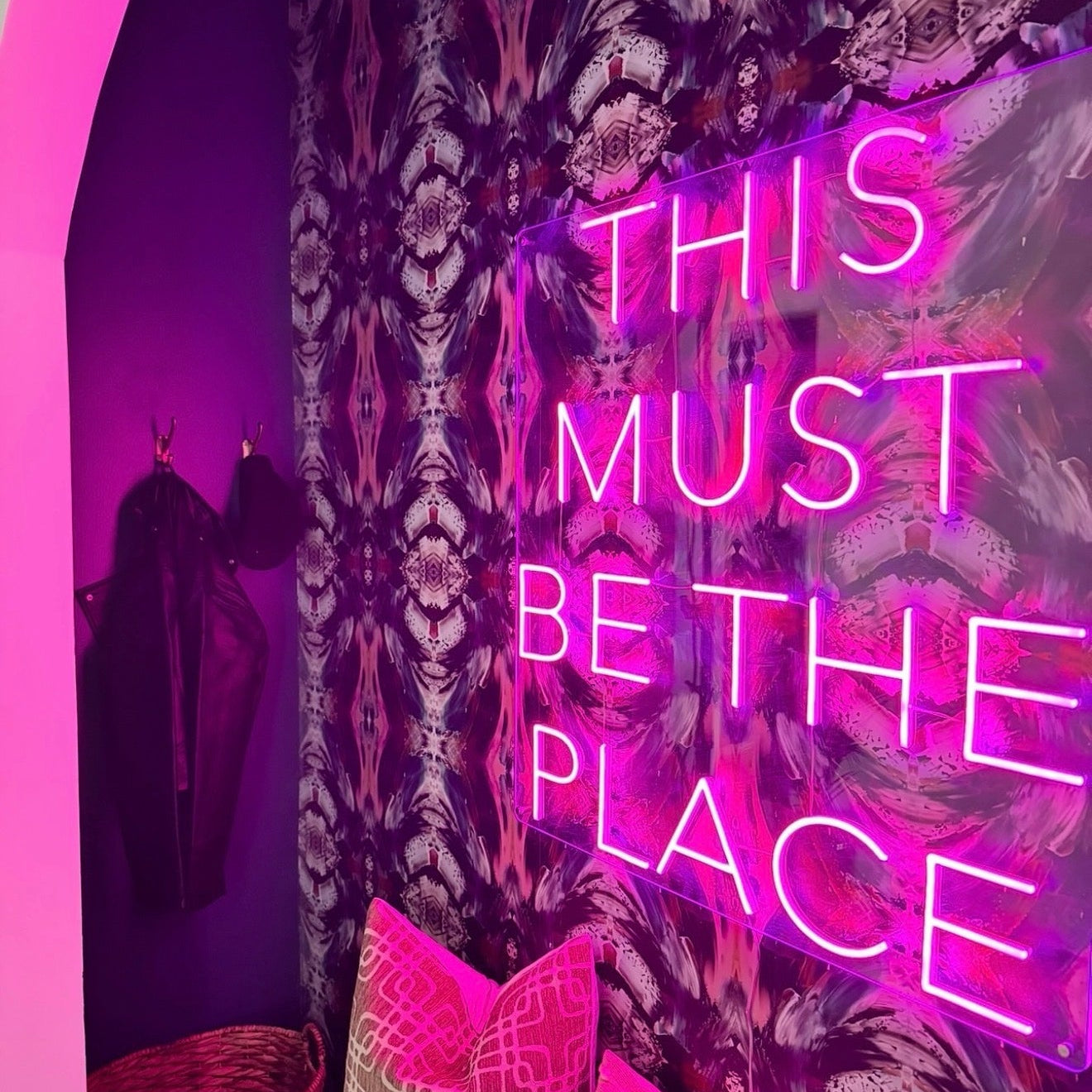 Neon sign reading 'THIS MUST BE THE PLACE' in a room with patterned wall and pink accents.