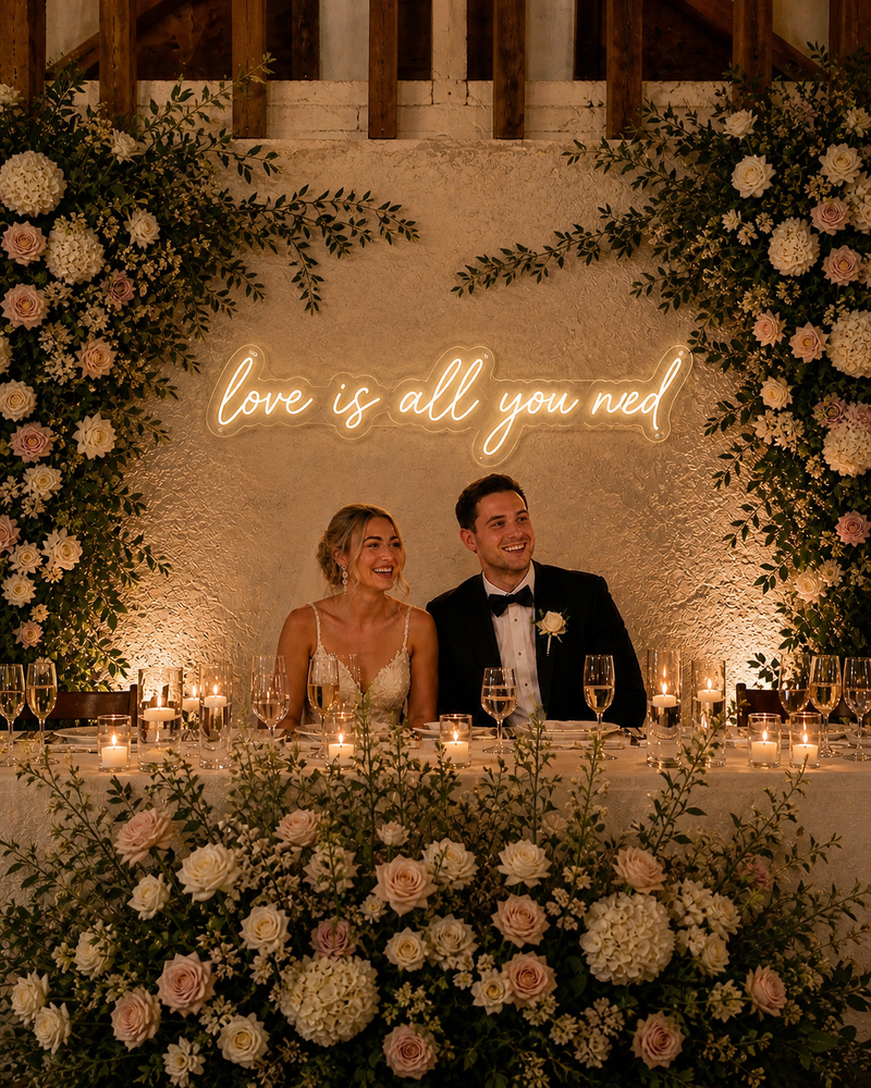 Couple sitting at a decorated wedding table with floral arch and 'love is all you need' sign.