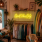 Neon sign 'Good Vibes Only' in a store with clothing racks and surfboards.
