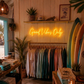 Neon sign reading 'Good Vibes Only' in a clothing store with surfboards and plants.