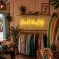 Neon sign 'Good Vibes Only' in a store with clothing racks and surfboards.