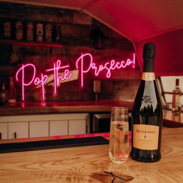 Bottle of Prosecco and a glass on a bar counter with a neon sign in the background.
