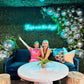 Two women sitting on a couch in a decorated room with disco balls and a neon sign.