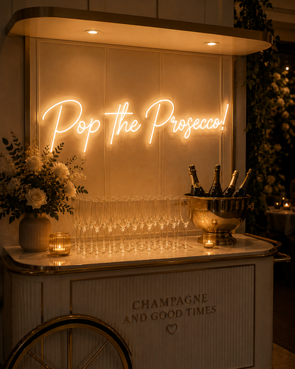 Decorative bar setup with 'Pop the Prosecco!' neon sign, champagne bottles, and glasses.
